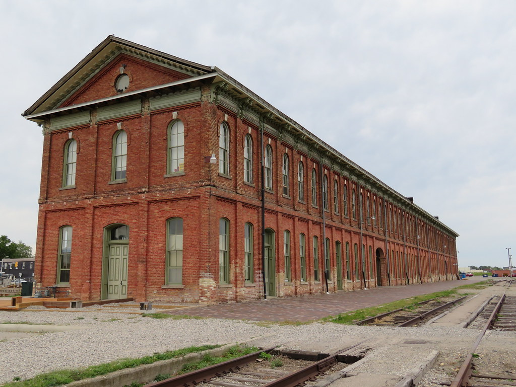 A reddish brown brick rectangular building with many windows, next to decaying railroad tracks 