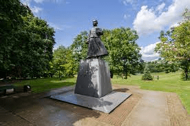 At the center of the image stands the large grey statue of Dr. Sun Yat-sen. Sun Yat-sen is standing on a large rectangular block and is holding a book. The statue is located in a large park surrounded by greenery. There is a large open space of grass in the background, along with many large green trees. Also in the upper background, you can see the blue sky, with a few white clouds. 