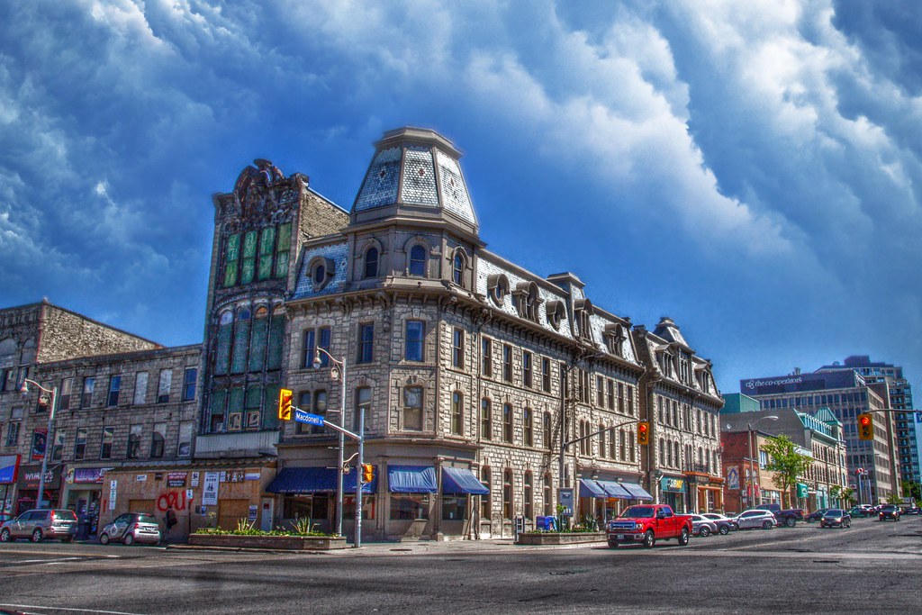 Street corner of downtown Guelph depicted with stone buildings and Petrie Building placed to the left. Main floor of Petrie Building covered for presumed renovations. Metal framing over front of building with numerous metal elements on top of building. Limescale noted over window, showing the building was left in disarray. 