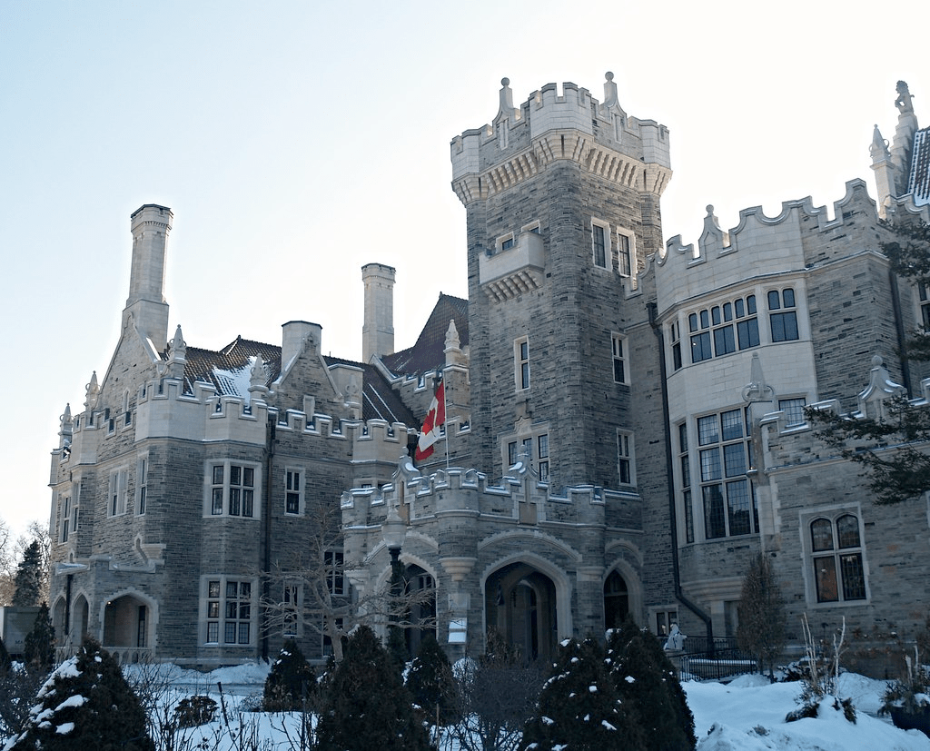 An old castle with a medieval look to it. The sun lights the back side of the castle with light grey stone and bricks that make up the structure. A Canadian flag waves proudly in the front of the stunning building. 
