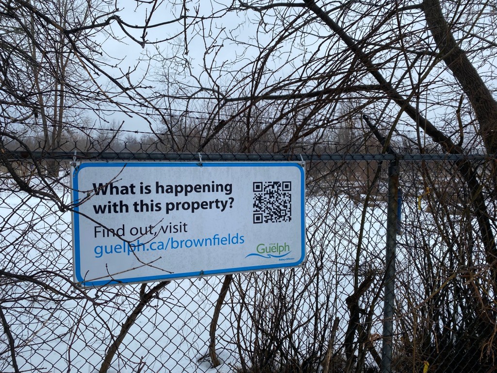 White sign hangs on a barbed wire fence. Sign says "What is happening with this property? Find out, visit guelph.ca/brownfields. City of Guelph." Land behind fence is covered in overgrown brush on an overcast day.
